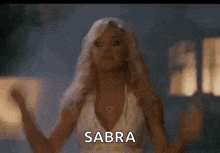 a woman in a yellow and white polka dot dress is screaming with her mouth open and the word sabra is on the screen .
