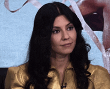 a woman with long black hair and a gold jacket looks at the camera