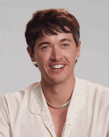 a man wearing a white shirt and a necklace smiles for the camera