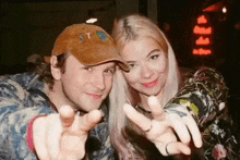 a man and a woman are posing for a picture and pointing at the camera . the man is wearing a hat .
