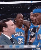 a man in a suit and tie is talking into a microphone while basketball players are standing around him .