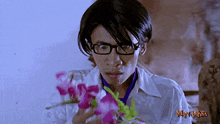a man wearing glasses is holding purple flowers in front of a man laksa sign