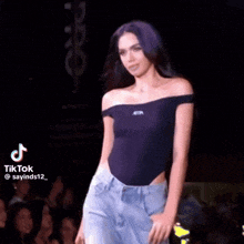 a woman in a black off the shoulder top and jeans is walking down the runway .