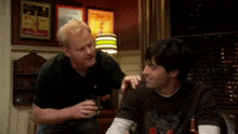 two men are sitting at a table talking to each other . one of the men is holding a glass of beer .
