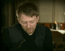 a man with a beard wearing a black vest is sitting on a couch .