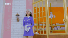 a woman in a purple dress and white hat is standing in front of a building .