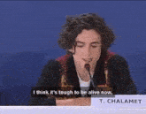 a young man is sitting at a table with a microphone and saying i think it is tough to be alive now .