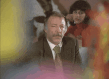 a man in a suit and tie sits in front of a woman in a red turtleneck