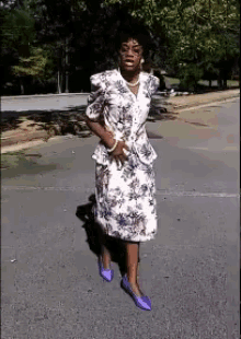 a woman in a floral dress and purple shoes stands on a street