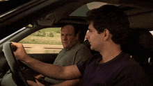 two men are sitting in a car one is driving and the other is sitting in the back seat
