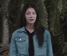 a woman in a denim jacket stands in front of a fern
