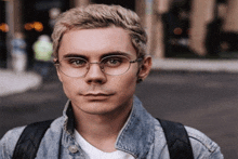 a young man wearing glasses and a denim jacket is looking at the camera