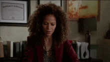 a woman with curly hair is sitting at a desk in front of a framed certificate of achievement .