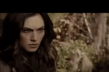 a woman with long dark hair is standing in the woods looking at the camera .