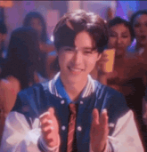 a young man in a baseball jacket and tie is clapping his hands in front of a crowd .