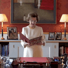 a woman sitting at a desk reading a book with a picture of a man on the wall in the background