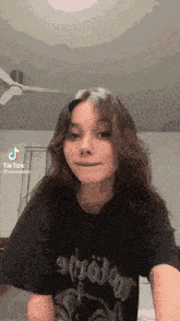 a girl wearing a black t-shirt is standing in front of a ceiling fan and looking at the camera .