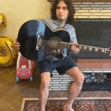 a man playing a guitar in front of a fireplace with a little tikes car in the background