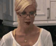 a woman wearing glasses is standing in a kitchen with a glass of wine in her hand .