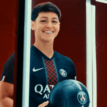 a young man wearing a qatar airways jersey smiles while holding a soccer ball
