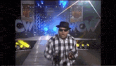 a man in a plaid shirt and fedora is walking down a runway .