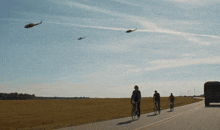 a group of people riding bicycles down a road with helicopters flying overhead