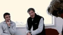 three men are sitting in front of a window and one is covering his mouth with his finger .