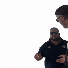 a man wearing sunglasses and a black adidas shirt talks to another man