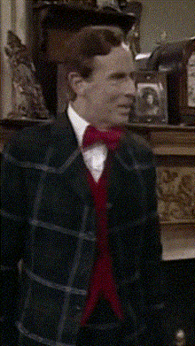 a man in a plaid suit and red bow tie stands in front of a fireplace
