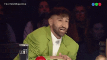 a man in a green jacket sits at a table with a cup that says talent on it