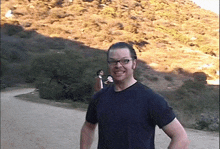 a man wearing glasses stands on a dirt road in front of a mountain