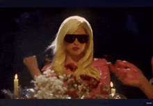 a woman in a blonde wig and sunglasses is sitting at a table with candles and flowers