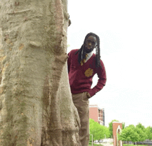 a man in a maroon sweater with a gold emblem on it stands next to a tree