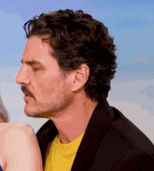 a man with curly hair and a beard is wearing a black jacket and a yellow shirt