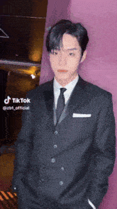 a man in a suit and tie is standing in front of a purple wall with his hands in his pockets .
