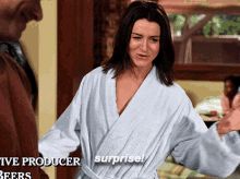 a woman in a bathrobe says surprise in front of a man in a brown shirt