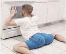 a man in blue shorts is kneeling on the floor looking into an oven .