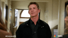 a man in a black jacket stands in a hallway with a nbc logo on the bottom