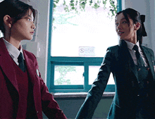 two girls in school uniforms are holding hands in front of a window with a no smoking sign on it