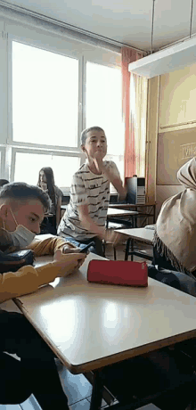 a boy wearing a mask is sitting at a desk in a classroom with a red pencil case