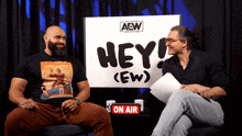 two men sitting in front of a sign that says hey