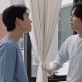 two men are standing next to each other in front of a window and looking at each other .