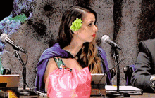 a woman with a flower in her hair sits at a table with microphones