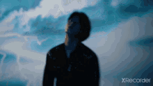 a man in a black jacket is standing in front of a blue sky with clouds in the background .