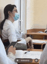 a woman wearing a mask sits at a desk in a classroom with a recording of her being played