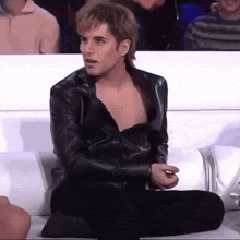 a man in a leather jacket is sitting on a couch with his legs crossed .