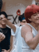 a man with red hair is wearing a white tank top and smiling while standing in front of a group of people .