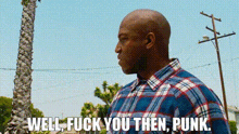 a man in a plaid shirt is standing in front of a palm tree and saying well fuck you then punk .