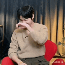 a man in a tan sweater is sitting in a red chair and covering his nose with his hand