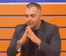 a man in a suit is sitting in front of a wall with his hands folded in prayer .